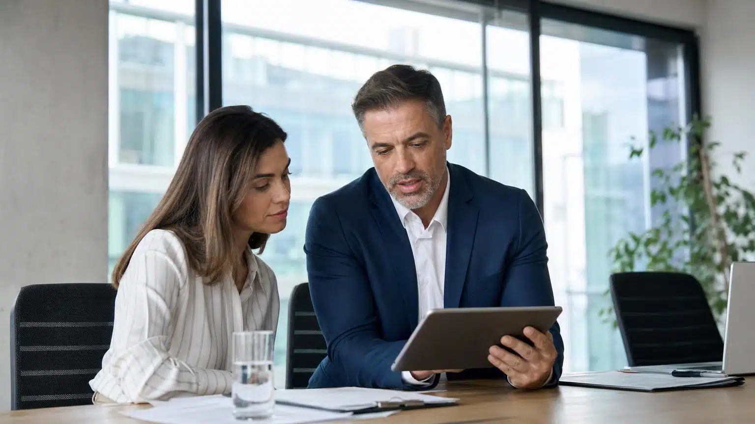 Two professionals in business attire review data on a digital tablet while sitting at a desk in a bright, modern office.