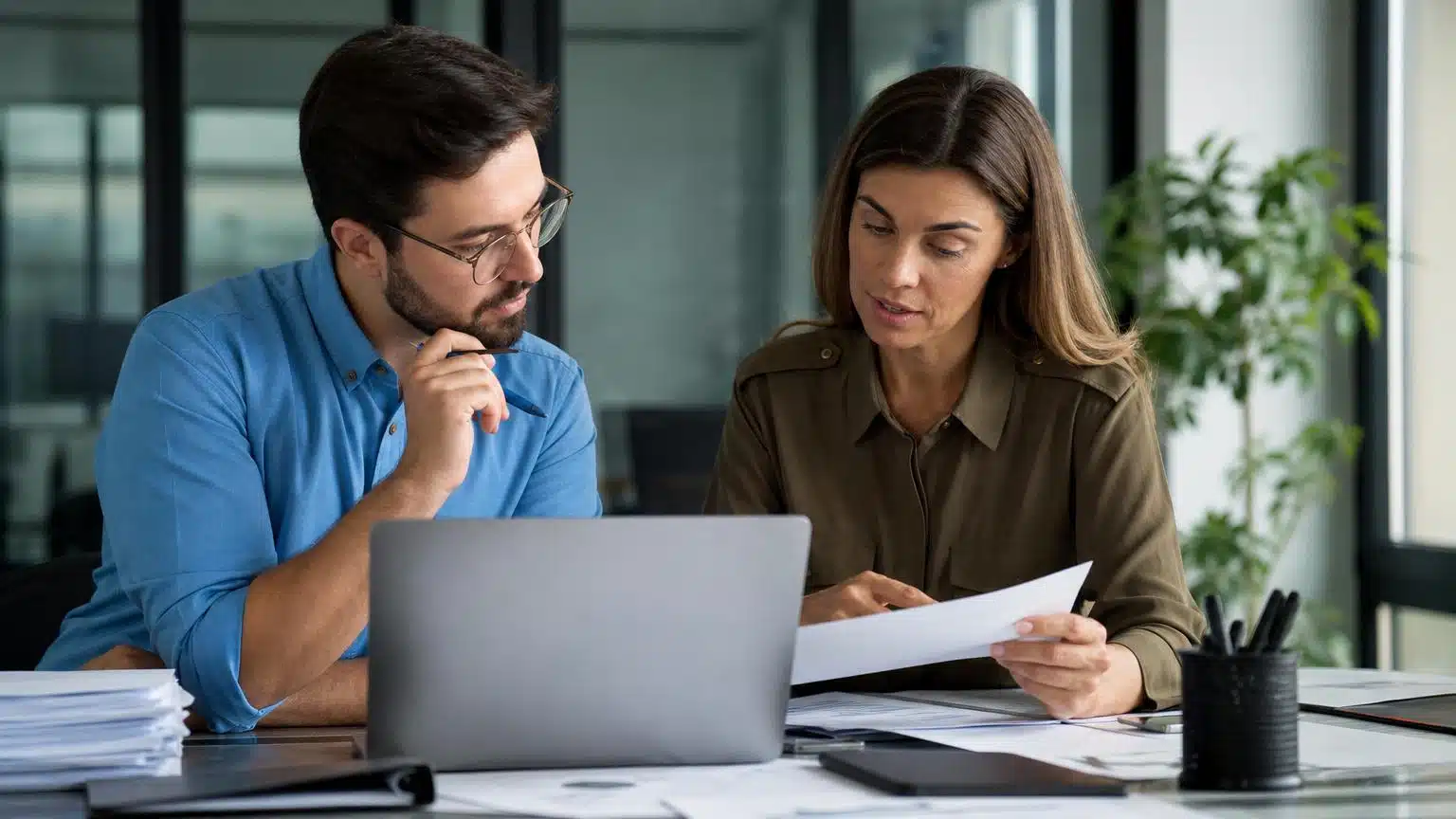 Two colleagues in a modern office reviewing paper documents and a laptop screen during a professional meeting.