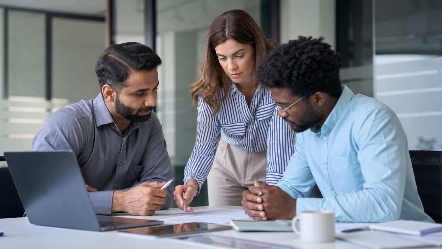 Three diverse professionals in a modern office review documents and a laptop together, focusing intently on a shared project at a white desk.
