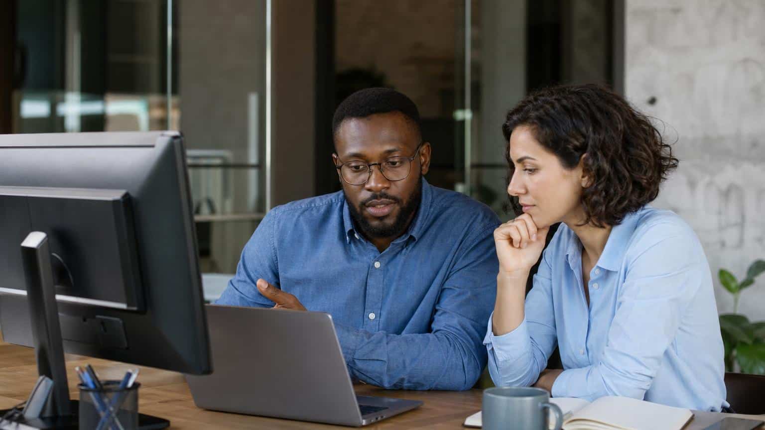 Two diverse professionals in a modern office focused on a laptop screen while reviewing data and collaborating on a project.
