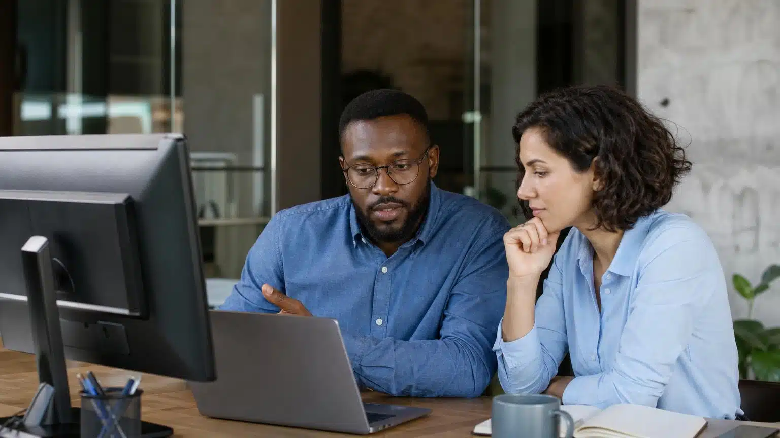 Two diverse professionals in a modern office focused on a laptop screen while reviewing data and collaborating on a project.